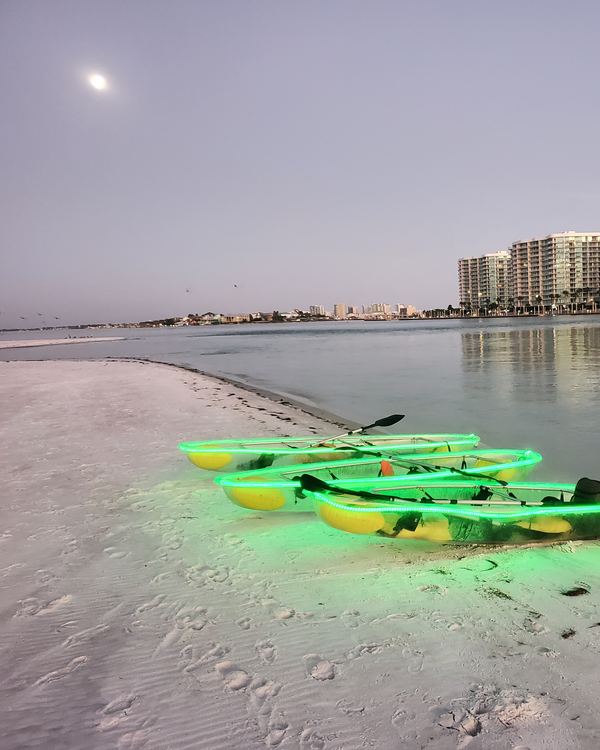 Glowing green kayaks pulled up on a white-sand beach at dusk, with the moon and the Orange Beach condos behind them