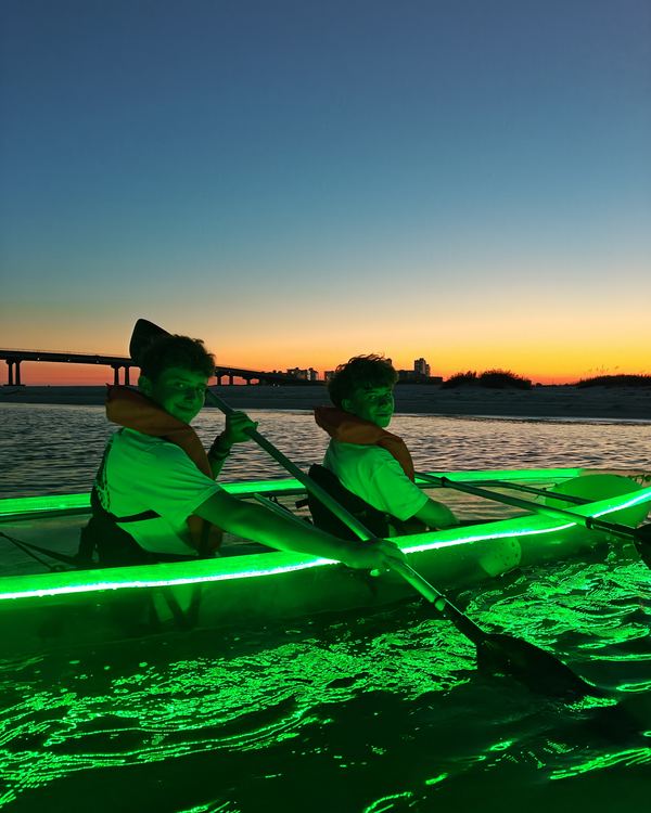 Two paddlers in a glowing clear kayak at sunset, with the bridge on the horizon