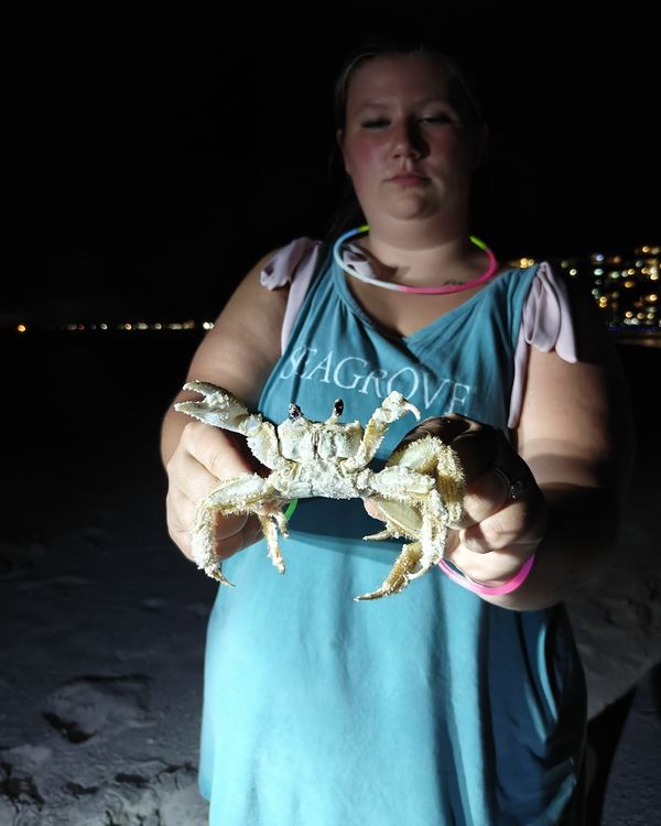 A guest in a teal top holding up a large crab with both hands on the beach at night