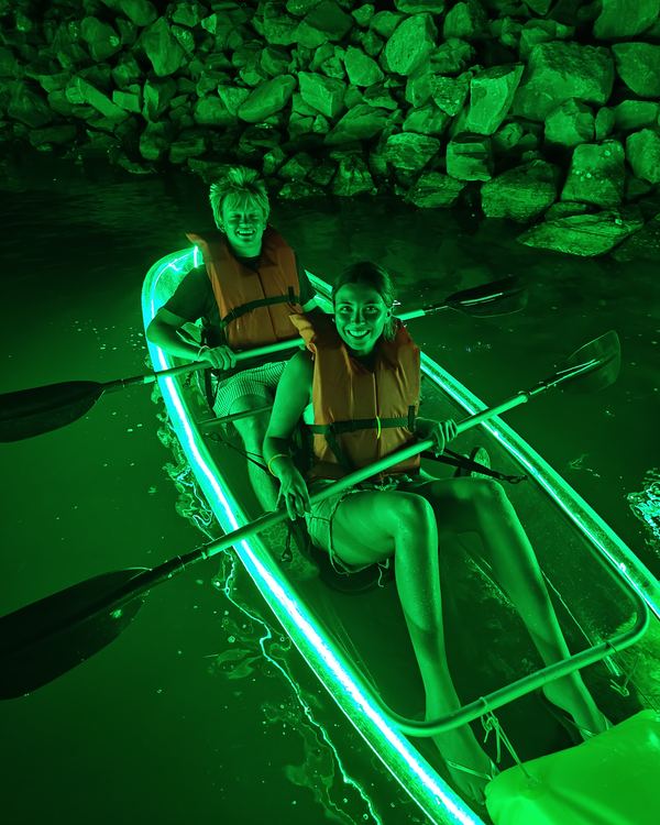Two smiling guests in life jackets paddling a clear kayak lit green beside the marina rocks