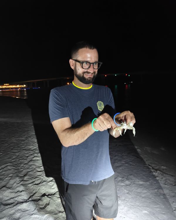 A smiling guest in glasses holding up a crab on the beach at night