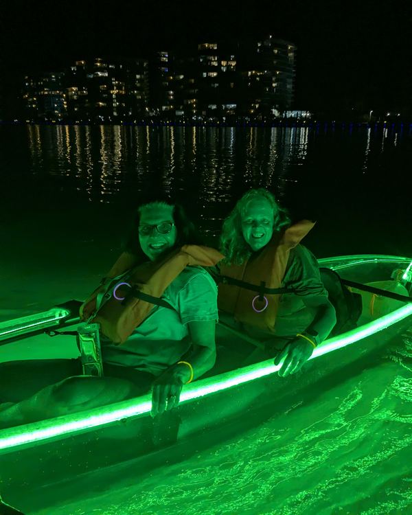 Two smiling guests in life jackets in a glowing kayak, with the lit condos across the bay behind them