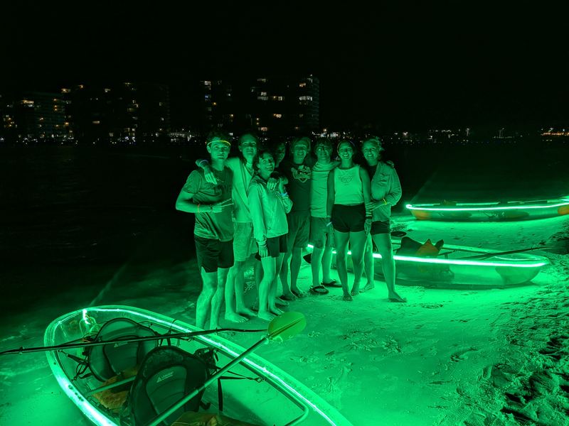 About eight guests standing together on the sand at night beside glowing clear kayaks, with the city lights across the water behind them