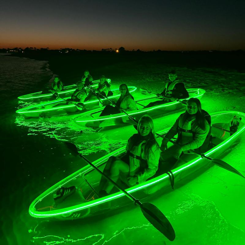 A group of guests in several green-glowing clear kayaks near the shore at dusk, with the last orange sunset glow on the horizon